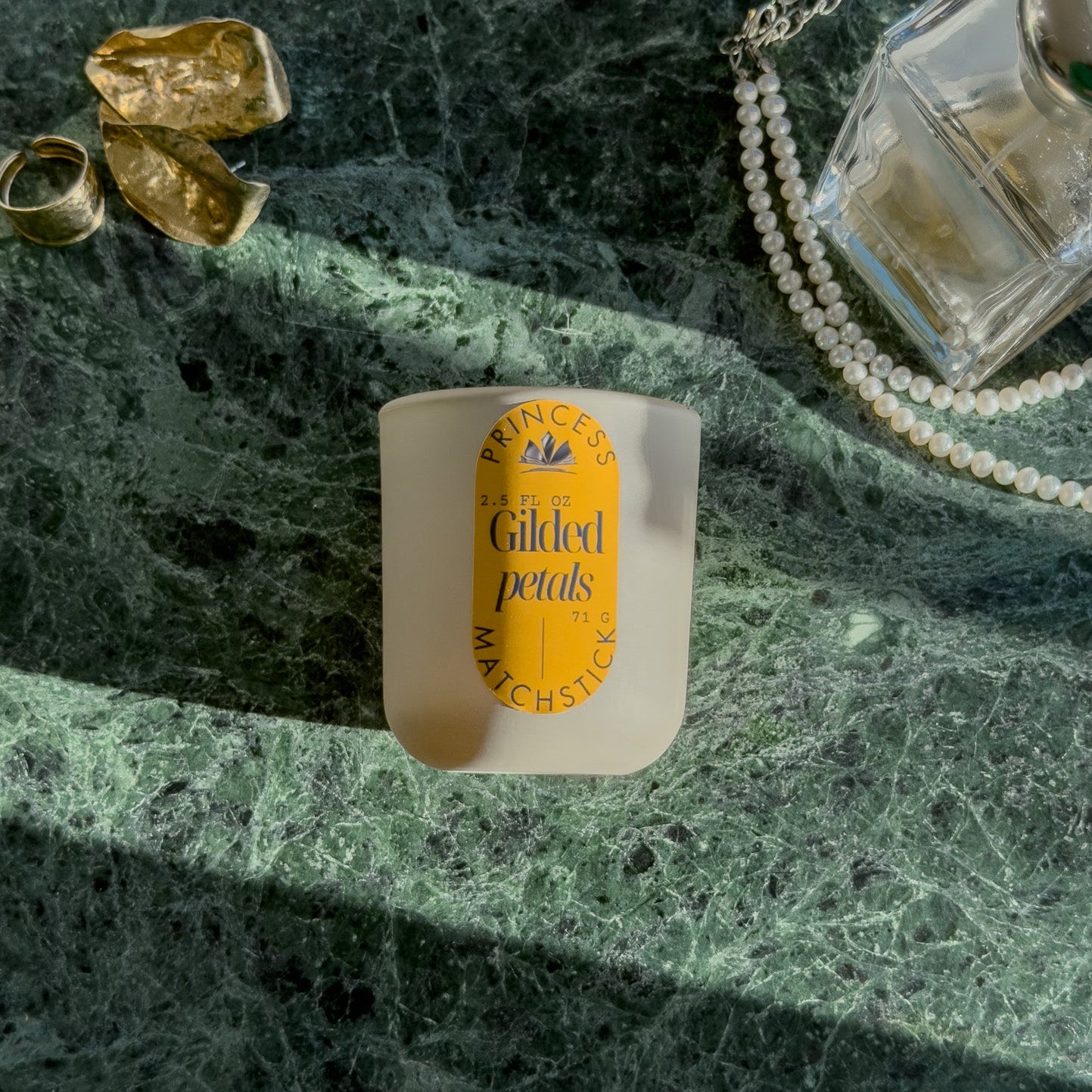 Small container with a yellow label on a green marble surface with jewelry and perfume bottle.